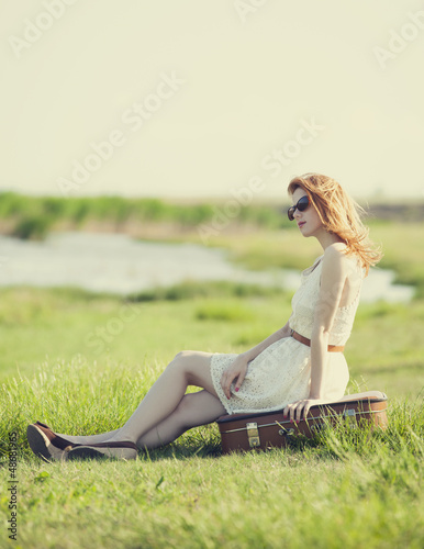 Photo: Young fashion girl with suitcase sitting at spring grass near la Young fashion girl with suitcase sitting at spring grass near la