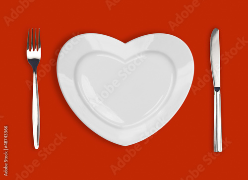 Photo: plate in shape of heart, table knife and fork on red background plate in shape of heart, table knife and fork on red background