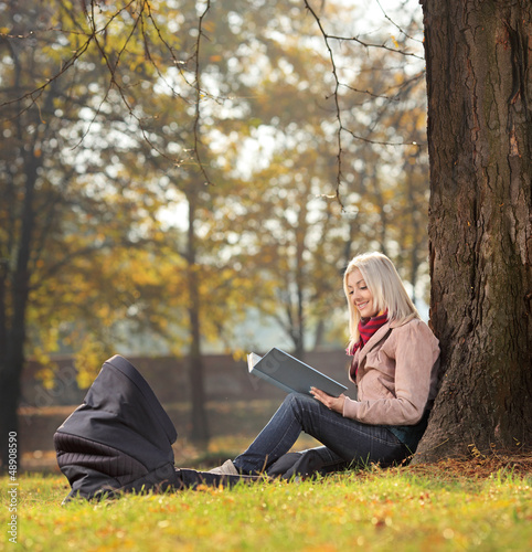Photo: Young mother sitting in a park reading a book with a baby in a c Young mother sitting in a park reading a book with a baby in a c