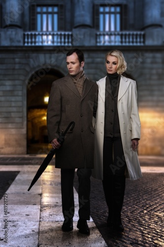 Photo: Elegant couple in coats against building facade in evening Elegant couple in coats against building facade in evening