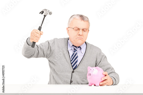 Photo: Angry mature man trying to break a piggy bank with a hammer Angry mature man trying to break a piggy bank with a hammer