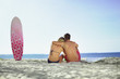 Photo: Rear view of a cute couple sitting on the beach near a surfboard Rear view of a cute couple sitting on the beach near a surfboard
