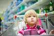 Photo: Adorable girl sit in shopping cart in supermarket with bottles o Adorable girl sit in shopping cart in supermarket with bottles o