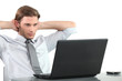 Young businessman at laptop with arms behind head.