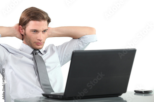 Photo: Young businessman at laptop with arms behind head. Young businessman at laptop with arms behind head.