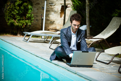 Photo: Young Businessman with Computer next to Swimming Pool Young Businessman with Computer next to Swimming Pool