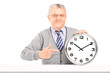 Foto: Mature man sitting and pointing on a wall clock Mature man sitting and pointing on a wall clock