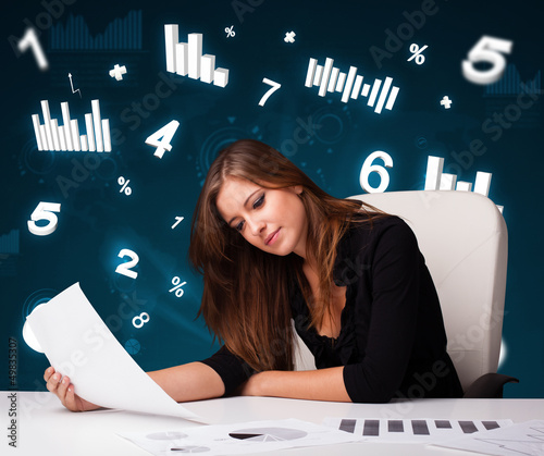 Photo: Young businesswoman sitting at desk with diagrams and statistics Young businesswoman sitting at desk with diagrams and statistics