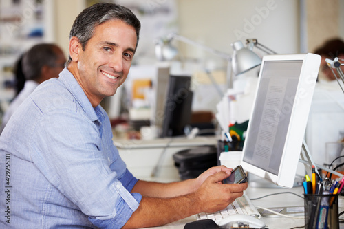 Photo: Man Using Mobile Phone At Desk In Busy Creative Office Man Using Mobile Phone At Desk In Busy Creative Office