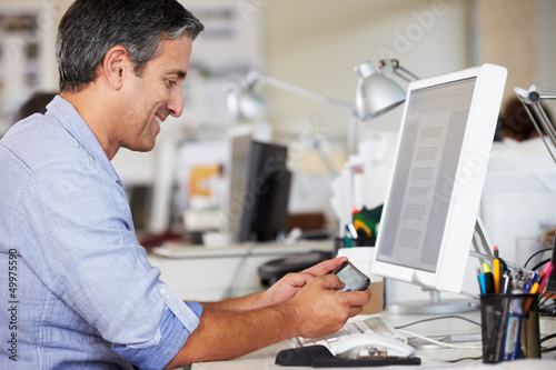 Photo: Man Using Mobile Phone At Desk In Busy Creative Office Man Using Mobile Phone At Desk In Busy Creative Office