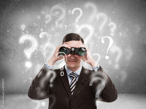 Photo: Handsome business man with binoculars and question marks above h Handsome business man with binoculars and question marks above h