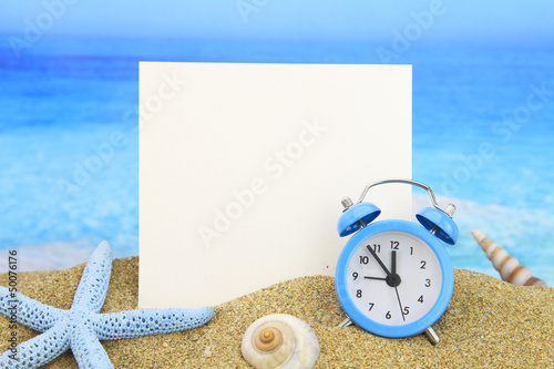 Photo: Summer time. Paper card and alarm clock on the beach Summer time. Paper card and alarm clock on the beach