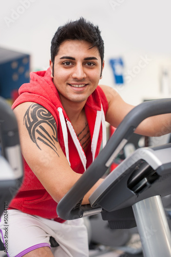 Photo: Young man training in the gym with bike. Young man training in the gym with bike.
