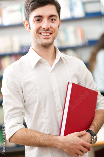 Photo: Portrait of a smiling male student holding a book in a library Portrait of a smiling male student holding a book in a library
