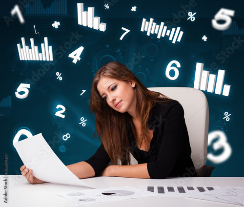 Photo: Young businesswoman sitting at desk with diagrams and statistics Young businesswoman sitting at desk with diagrams and statistics