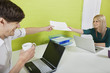Photo: Happy young businesspeople passing documents to each other with laptops at desk Happy young businesspeople passing documents to each other with laptops at desk