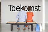 Photo: Rear view of couple seated on bench reading Dutch text "Toekomst" (future) on wall Rear view of couple seated on bench reading Dutch text "Toekomst" (future) on wall