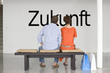 Photo: Rear view of couple seated on bench reading German text "Zukunft" (future) on wall Rear view of couple seated on bench reading German text "Zukunft" (future) on wall
