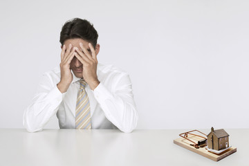 Photo: Worried businessman at table model home in mouse trap representing expensive real estate costs Worried businessman at table model home in mouse trap representing expensive real estate costs