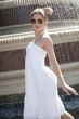 Photo: Side view of happy young woman in white halter neck dress standing by water fountain Side view of happy young woman in white halter neck dress standing by water fountain