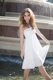 Photo: Portrait of happy young woman in white halter neck dress standing by water fountain Portrait of happy young woman in white halter neck dress standing by water fountain