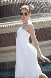 Photo: Side view of happy young woman in white halter neck dress standing by water fountain Side view of happy young woman in white halter neck dress standing by water fountain