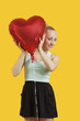 Photo: Portrait of beautiful young woman peeking through heart shaped balloon over yellow background Portrait of beautiful young woman peeking through heart shaped balloon over yellow background