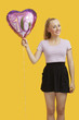 Photo: Portrait of beautiful young woman holding heart shaped birthday balloon over yellow background Portrait of beautiful young woman holding heart shaped birthday balloon over yellow background