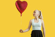 Photo: Beautiful young woman looking at heart shaped balloon over yellow background Beautiful young woman looking at heart shaped balloon over yellow background