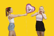 Photo: Happy young woman giving birthday balloon to amazed woman standing over yellow background Happy young woman giving birthday balloon to amazed woman standing over yellow background