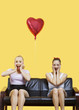 Photo: Portrait of two amazed young women sitting on sofa with heart shaped balloon over yellow background Portrait of two amazed young women sitting on sofa with heart shaped balloon over yellow background