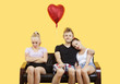 Photo: Portrait of young couple sitting on sofa with female friend feeling left out over yellow background Portrait of young couple sitting on sofa with female friend feeling left out over yellow background