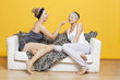 Photo: Happy young woman applying face pack on friend's face while sitting on sofa against yellow wall Happy young woman applying face pack on friend's face while sitting on sofa against yellow wall