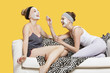 Photo: Two happy young women applying face pack while sitting on sofa over yellow background Two happy young women applying face pack while sitting on sofa over yellow background