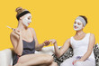 Photo: Young woman with face pack filing friend's nails while sitting on sofa over yellow background Young woman with face pack filing friend's nails while sitting on sofa over yellow background