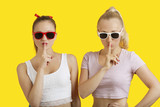 Photo: Portrait of two playful young women with fingers on lips over yellow background Portrait of two playful young women with fingers on lips over yellow background