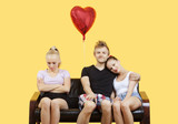 Photo: Portrait of young couple sitting on sofa with female friend feeling left out over yellow background Portrait of young couple sitting on sofa with female friend feeling left out over yellow background