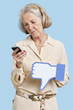 Foto: Senior woman with cell phone holding fake dislike button against blue background Senior woman with cell phone holding fake dislike button against blue background