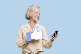 Photo : Senior woman with cell phone holding fake like button against blue background Senior woman with cell phone holding fake like button against blue background