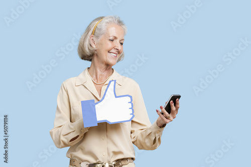Photo: Senior woman with cell phone holding fake like button against blue background Senior woman with cell phone holding fake like button against blue background
