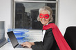 Photo: Portrait of senior businesswoman in superhero costume using laptop at office desk Portrait of senior businesswoman in superhero costume using laptop at office desk