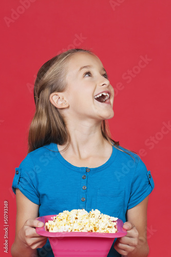 Photo: Portrait of surprised young girl holding popcorn container against red background Portrait of surprised young girl holding popcorn container against red background