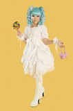 Photo: Full length portrait of young woman dressed as a doll holding lollipop over yellow background Full length portrait of young woman dressed as a doll holding lollipop over yellow background