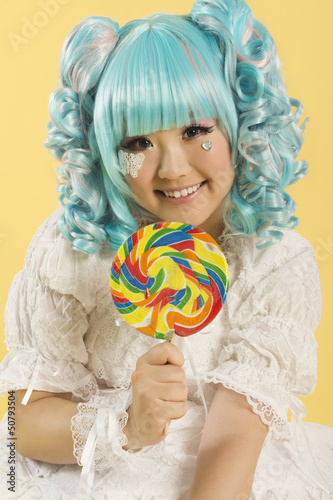 Photo: Portrait of smiling young woman dressed as a doll holding lollipop over yellow background Portrait of smiling young woman dressed as a doll holding lollipop over yellow background