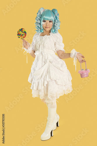 Photo: Full length portrait of young woman dressed as a doll holding lollipop over yellow background Full length portrait of young woman dressed as a doll holding lollipop over yellow background