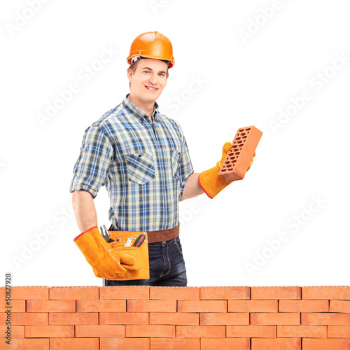 Photo: Male manual worker with helmet holding a brick behind a brick wa Male manual worker with helmet holding a brick behind a brick wa