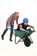 Two female construction workers racing in a wheelbarrow.