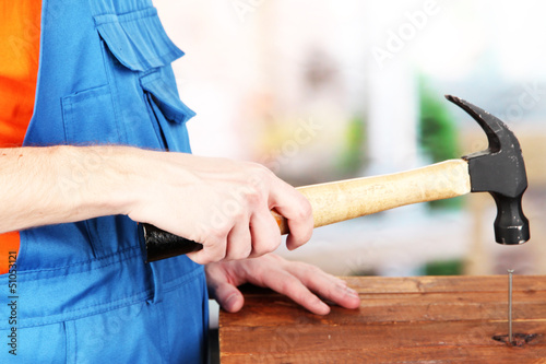 Photo: Builder hammering nails into board on room background Builder hammering nails into board on room background