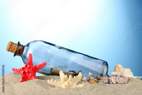 Photo: Glass bottle with note inside on sand, on blue background Glass bottle with note inside on sand, on blue background