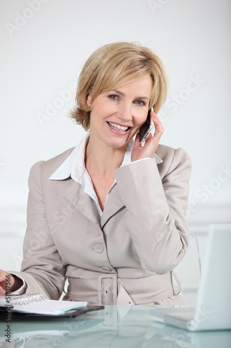 Photo: Businesswoman sat at desk holding mobile telephone Businesswoman sat at desk holding mobile telephone